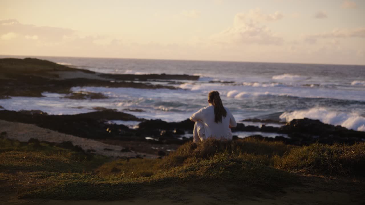A man stands on a grassy bluff gazing at the ocean waves rolling against rugged rocks, preparing to sit as the warm sunset light fills the horizon