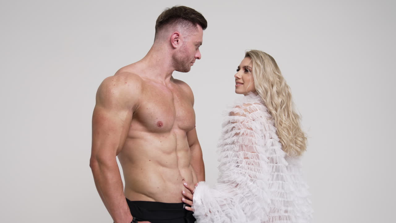 Beautiful long-haired blonde lady comes up to athletic man with naked torso. Woman walks around the man and the couple kissing. White backdrop.