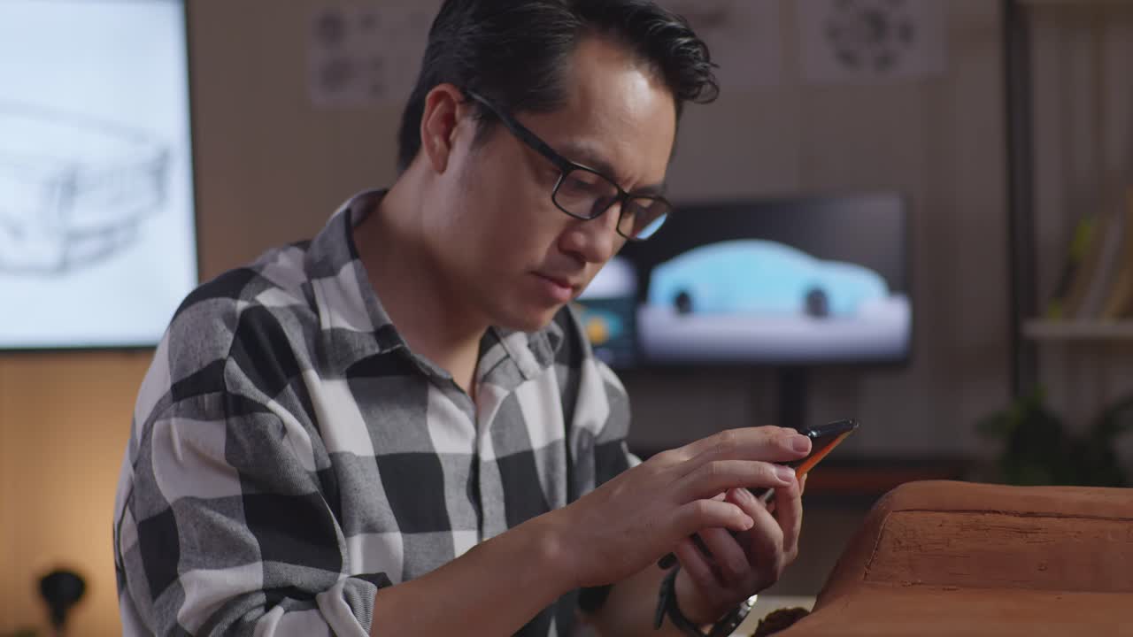 아시안 남성 자동차 디자이너가 스튜디오에서 스파라로 자동차 점토 조각을 작업하는 동안 스마트폰의 사진과 디자인을 비교합니다.