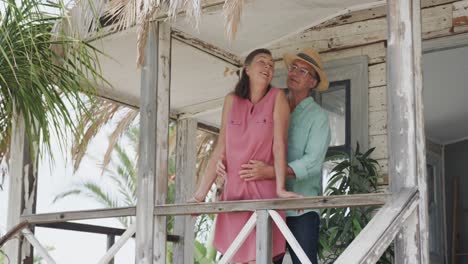Happy-senior-caucasian-couple-embracing-on-porch-of-wooden-beach-house,-in-slow-motion