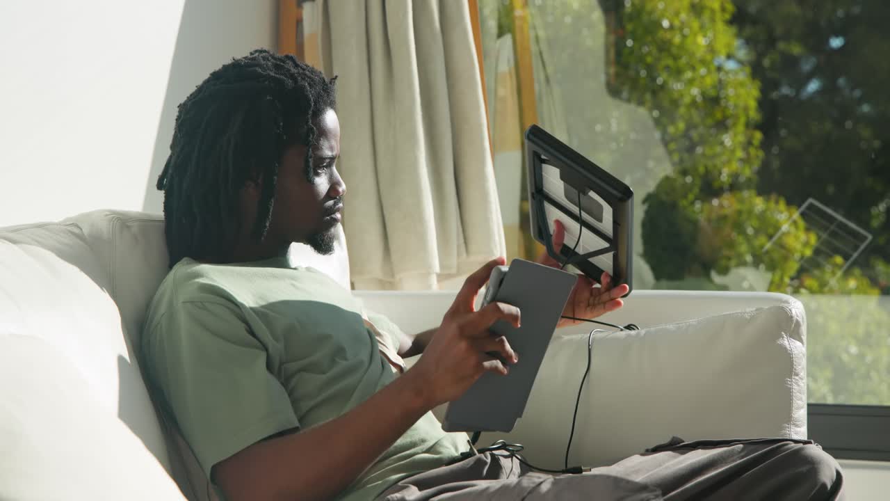 African American man on sofa reacting to sun, angling solar panel to window to charge tablet