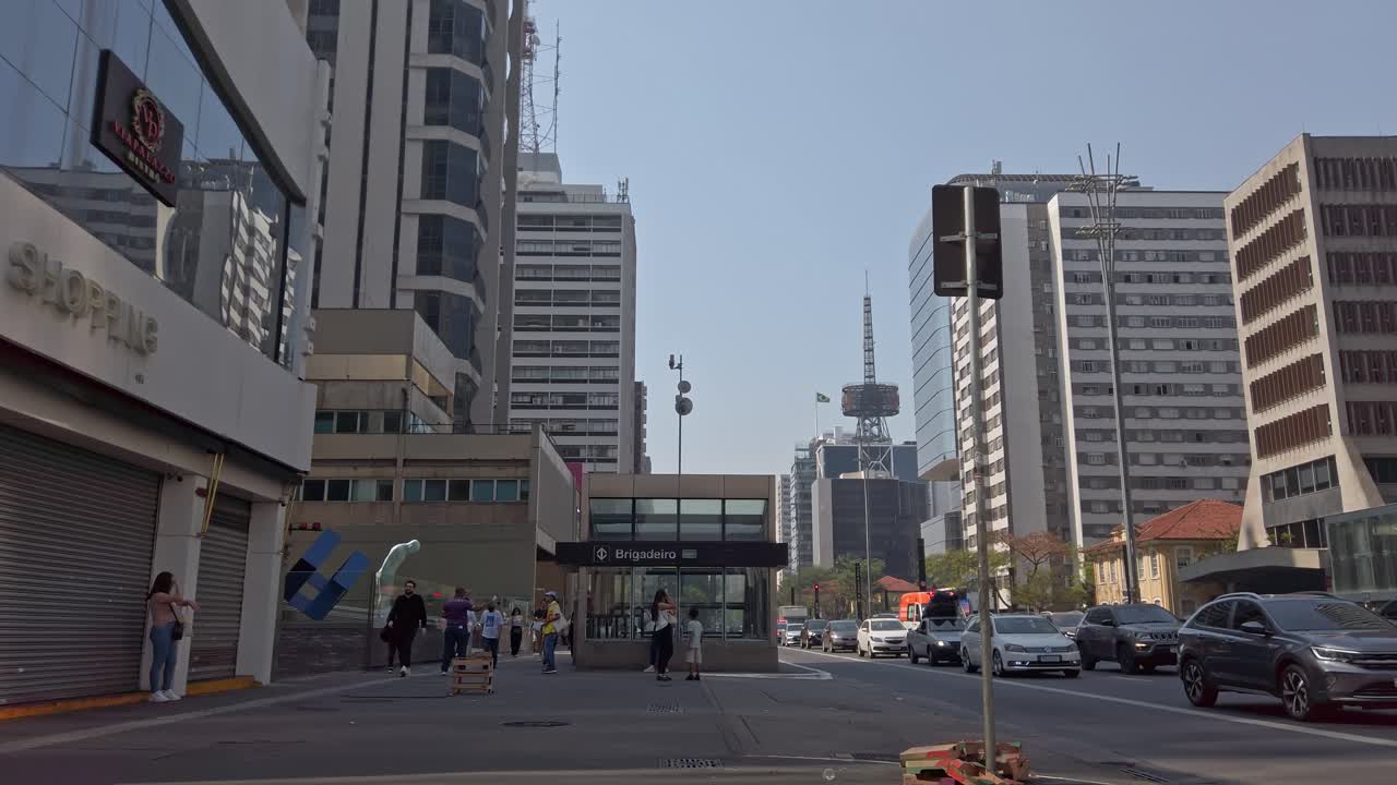 In São Paulo, locals and tourists navigate a lively street surrounded by tall buildings and clear blue skies. Cars drive by as people chat and walk