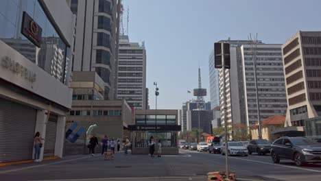 in são paulo, locals and tourists navigate a lively street surrounded by tall buildings and clear blue skies. cars drive by as people chat and walk
