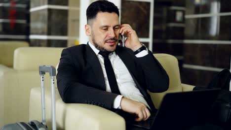 tilt up of confident businessman talking mobile phone and using laptop computer while sits on armchair in luxury hotel with luggage near him. travel, business and people concept