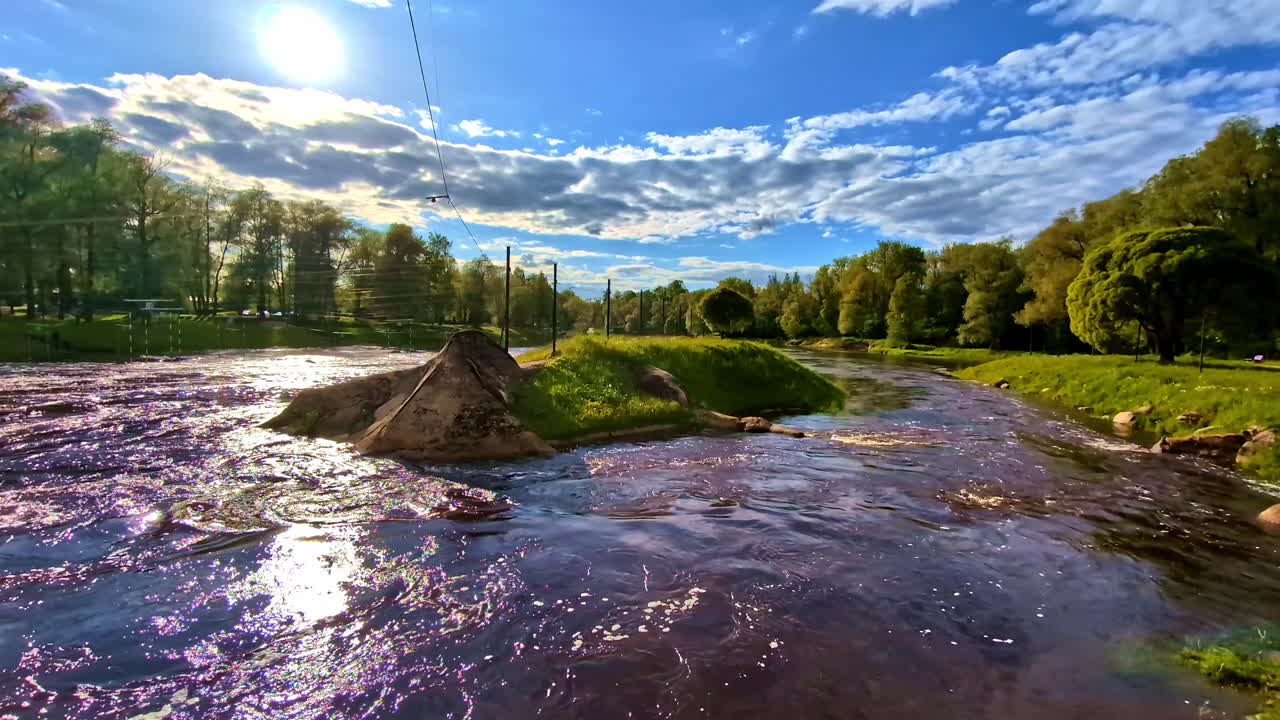 Scenic wide Gauja river bend with rapids, lush green banks and forest under cloudy blue sky in Valmiera, Latvia