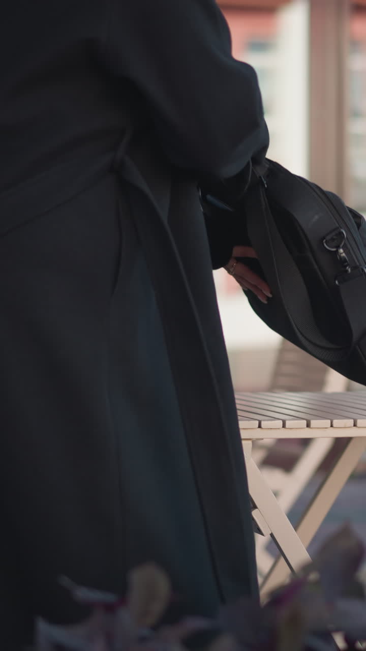 mujer se sienta en un café, coloca su bolsa en la mesa, y la abre, revelando su muslo, con un fondo urbano con edificios y una bolsa cercana