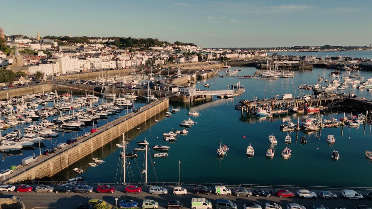 Drönarflygning över St Peter Port hamn, Guernsey, över sjövall, Albert Marina, Victoria Marina och fiskarhamnarna i sen eftermiddagssol, med staden och Belle Greve Bay i bakgrunden på avstånd.