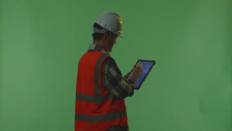 back view of asian male engineer with safety helmet using a tablet drawing the wind turbine blueprint while standing in the green screen background studio, during sunset or sunrise time