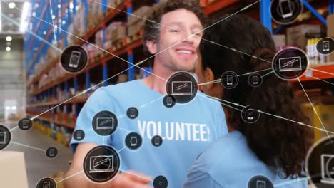 Animation-of-network-of-digital-icons-on-two-diverse-male-and-female-volunteers-hugging-at-warehouse