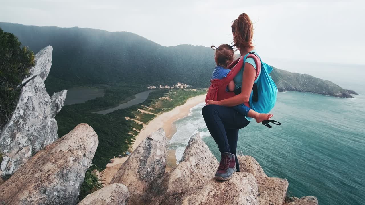 familia de senderismo en mal tiempo. madre de pie con el bebé en envoltura de correa en la cima de la montaña con magnífica vista del valle durante el día de viento y lluvia