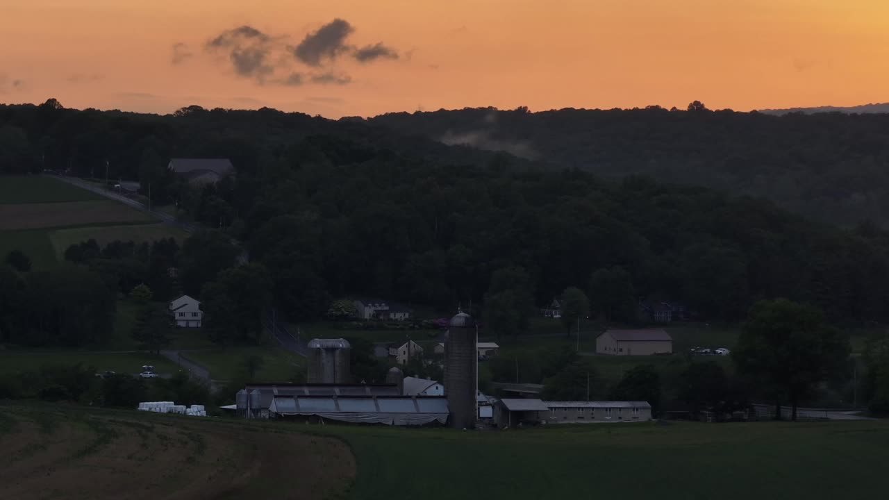 Aerial wide shot of american farm with silo storage during golden sunrise. Yellow lighting sky behind mountain and hills of America. Dusk scene in rural district of city.