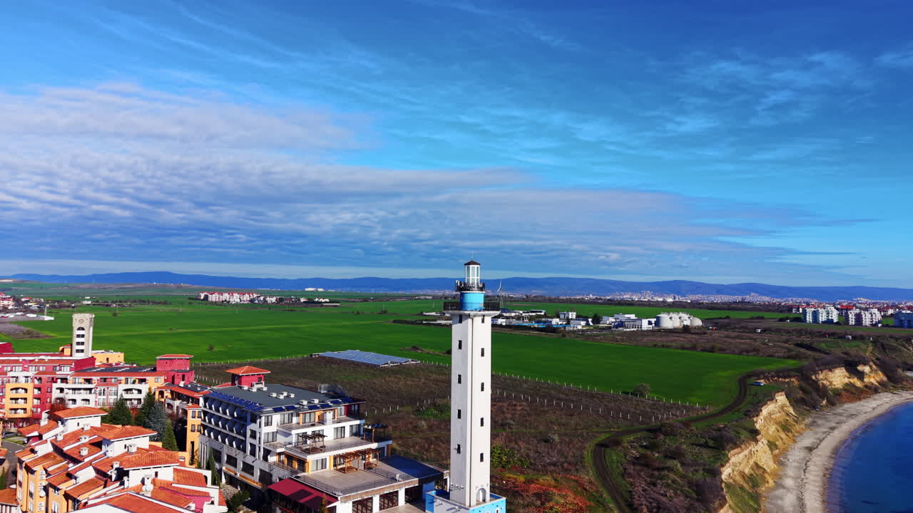 Vista aerea di un faro in cemento bianco situato tra un edificio residenziale e vasti campi agricoli verdi. Alto faro bianco vicino a campi verdi e alla costa.