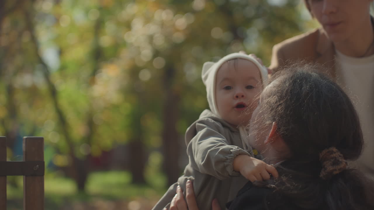 Nærbillede af bedstemor, der justerer babyhat i solrig park, mens far ser på, blid berøring og detaljer om strikhue, ømt omsorgsudveksling, der fremhæver familiens varme og tværgenerationelle bånd.