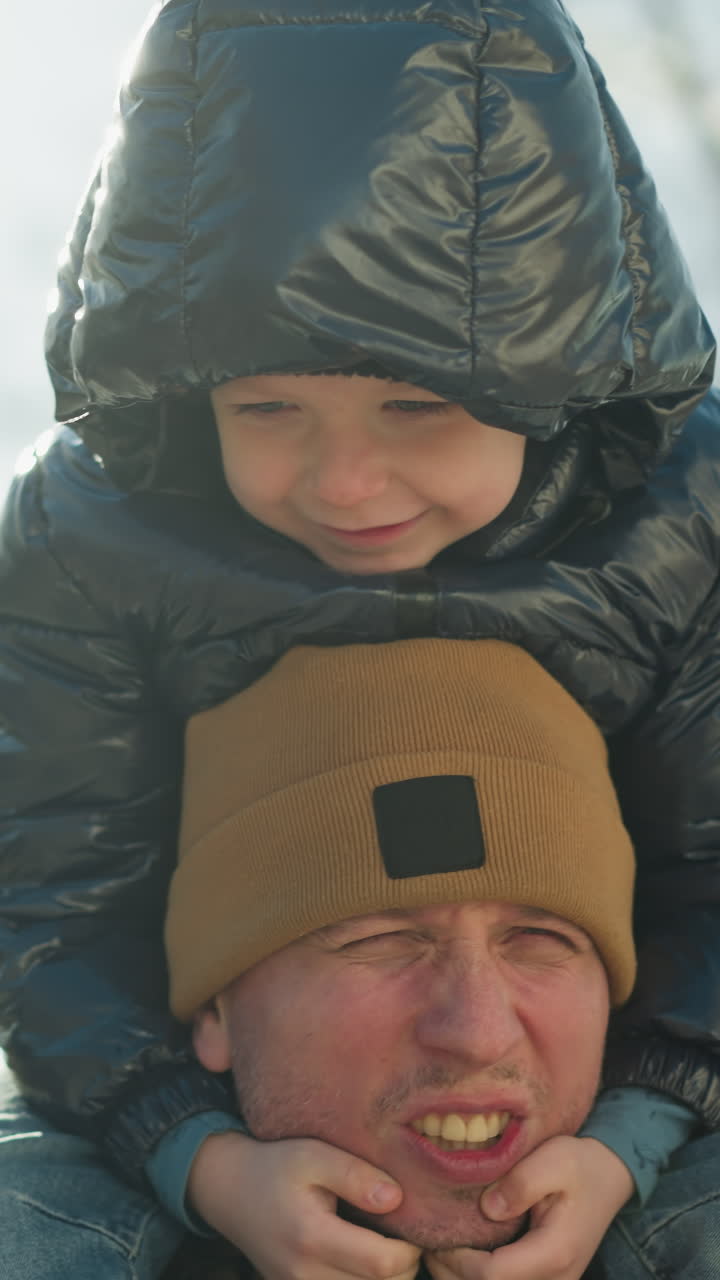 웃는 아이를 어깨에 고 있는 아버지의 클로즈업, 둘 다 기쁜 순간을 즐기고, 아이의 손은 아버지의 얼굴 에 앉아 있으며, 부드러운 빛의 광선이 뒤에서 반사됩니다.