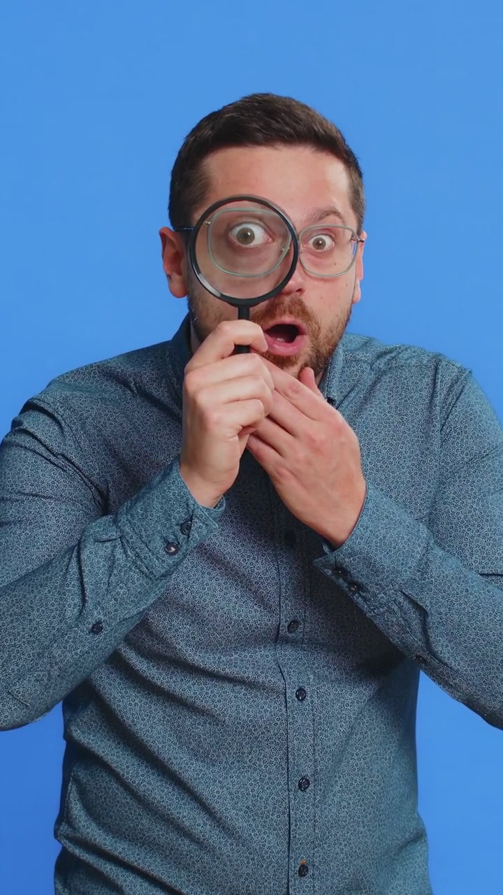 Hombre de mediana edad sostiene una lupa cerca de la cara, mirando a la cámara con un ojo grande y ampliado, analizando