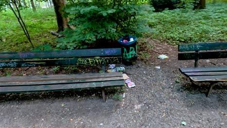 camera slowly pans past a weathered wooden bench with graffiti and a nearby trash bin in a leafy berlin park, under diffuse daylight