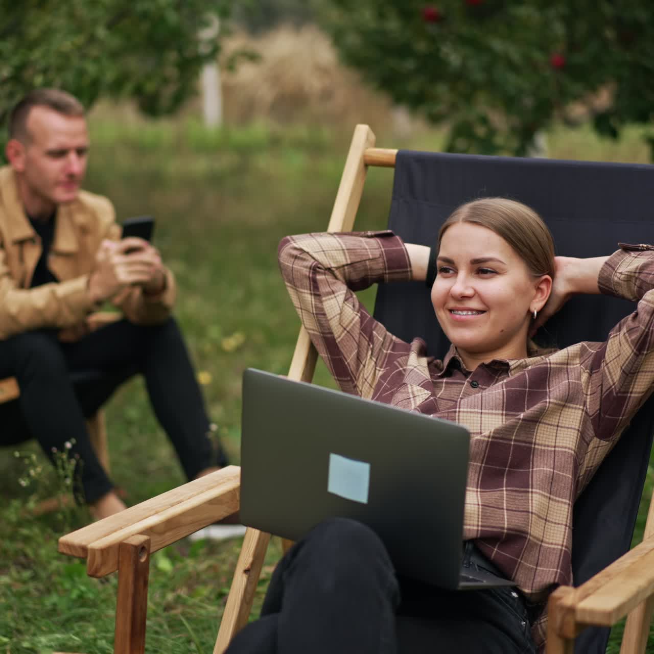 Kobieta i mężczyzna siedzą na krzesłach ogrodowych na łonie natury. Kobieta robi sobie przerwę od pracy na laptopie, a mężczyzna korzysta ze smartfona. W tle sad jabłoni