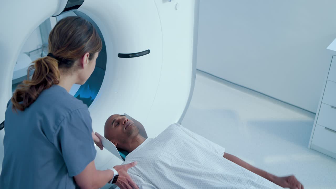 Paziente afroamericano in camice che ascolta un tecnico asiatico spiegare la scansione, disteso all'interno dello scanner