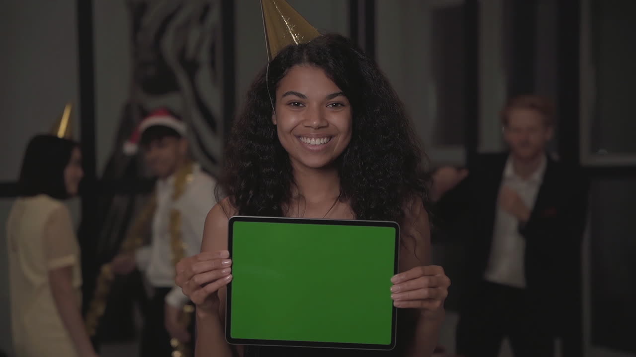 joven mujer negra feliz sosteniendo una tableta con pantalla verde maqueta y mirando a la cámara en la fiesta de fin de año