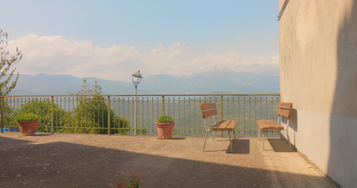 Terrazza silenziosa con panchine di legno vuote, piante in vaso e una valle montana nebbiosa oltre una ringhiera in metallo sotto una luce soffusa