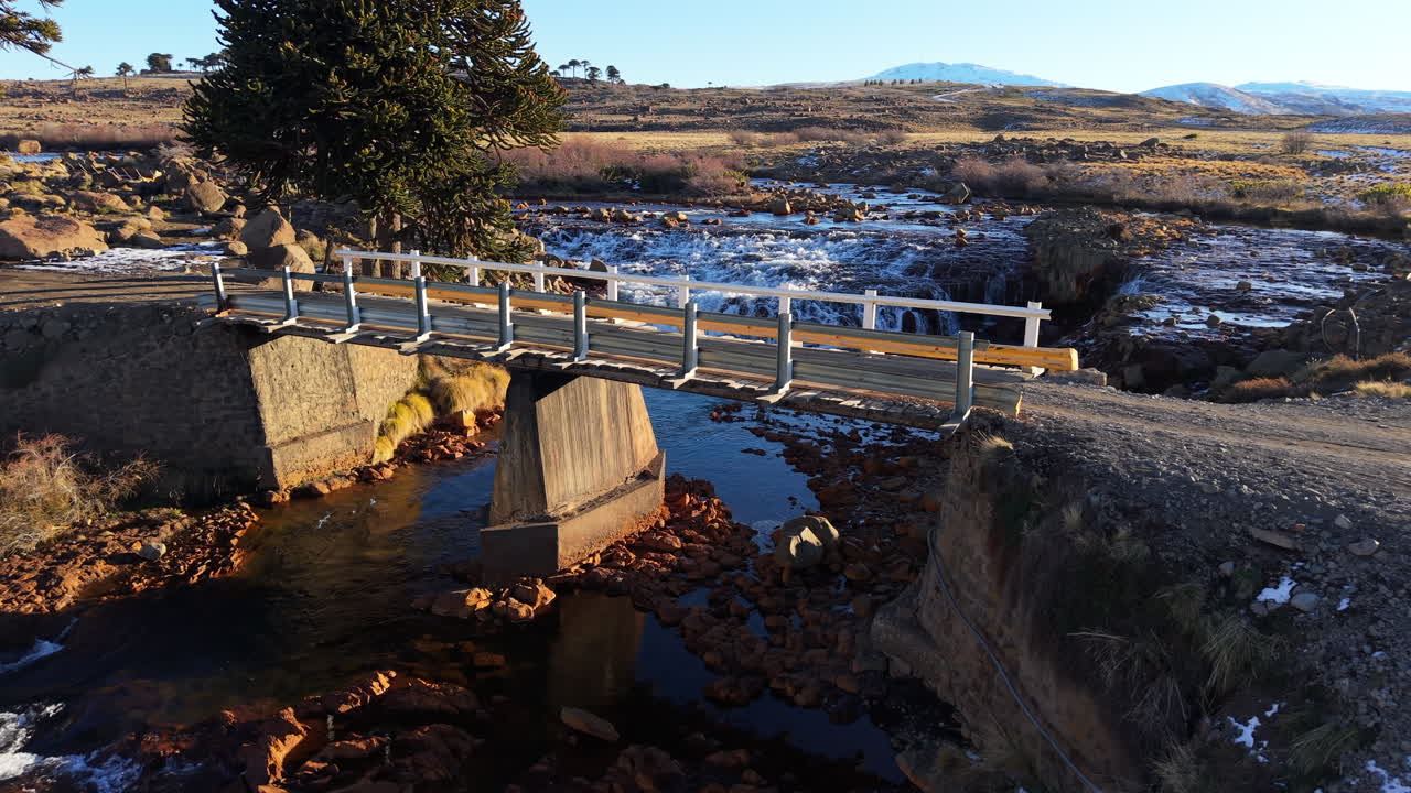 Elevated view of a small wooden bridge crossing the Rio Agrio above gentle cascades, framed by Andean steppe and volcanic terrain near Caviahue, Neuquén, in soft morning light