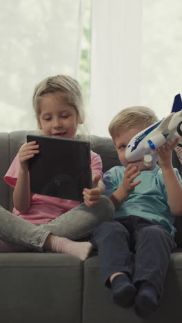 sister shows funny videos to little brother turning tablet while sitting on sofa. children use gadget and play with airplane toy in living room