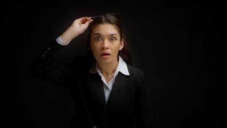 una mujer de negocios conmocionada levanta sus gafas de sorpresa contra un fondo negro.