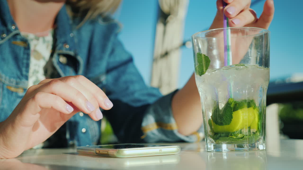 한 여성이 카페에서 스마트폰을 사용하고 있습니다. 옆에 칵테일 한 잔이 있어 휴식을 취하고 있습니다.