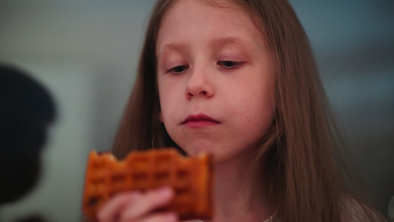 Close up of little girl joyfully savoring golden waffle, eyes closed in delight, wearing white shirt, with partial view of another person nearby against soft evening indoor background