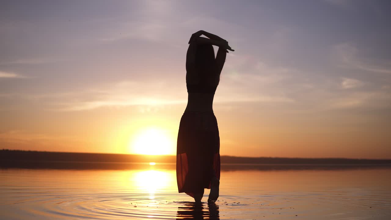 Silhouette of a slender girl walking on the water in yoga pants, raises her hands. Carefree, freedom. Incredible sunset in the background