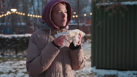 joven de pie en el parque comiendo maíz, limpiándose la boca con una servilleta, envuelta en plástico, escenario invernal al aire libre con suelo cubierto de nieve, luces bokeh en el fondo