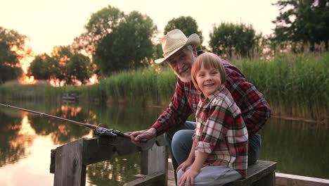 vista ravvicinata di un nonno e di suo nipote che pescano e parlano seduti sul molo del lago in una giornata estiva al tramonto