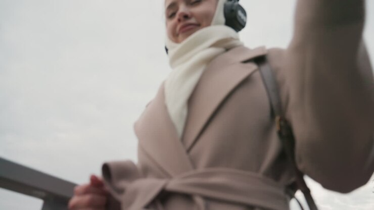 Donna che balla all'aperto. Donna che si muove gioiosamente lungo il lungomare. Signora che si muove a ritmo di musica sullo sfondo dello skyline. Figura femminile con sciarpa e stivali, vividamente espressiva durante una vivace danza all'aperto.