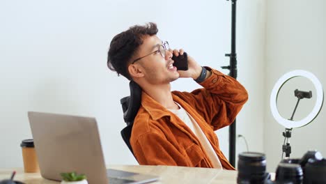 young male photographer with glasses talks on smartphone while sitting at desk with equipment and laptop