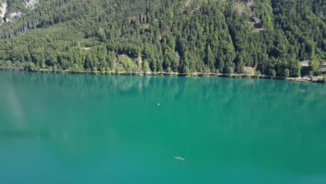teal coloured lake with grey rocks along the shoreline small boat floating on the waters view of the shore filled with multitude of deep green trees vivid palette mirror effect on the water