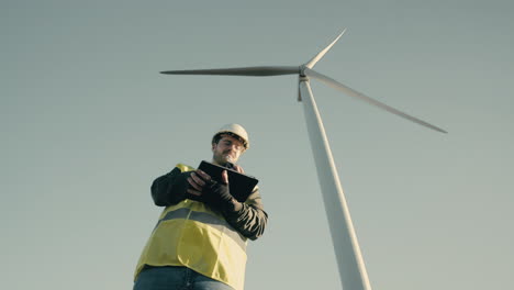 a professional caucasian engineer in a reflective vest and white helmet uses a tablet to audit wind turbines in a field, symbolizing the growth of renewable energy