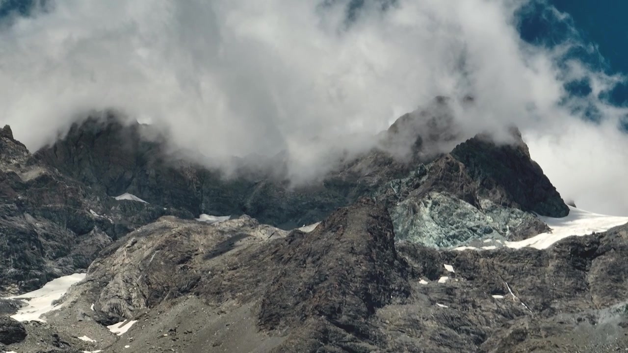 Nærbillede af dramatiske alpine bjergtoppe indhyllet i rullende skyer, der afslører dvælende gletsjerpletter mellem barske tinder under skiftende bjergvejr.