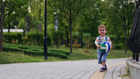 lovely brown-haired with a ball in hands stands in the park. kid walks by the paved road carrying a ball.