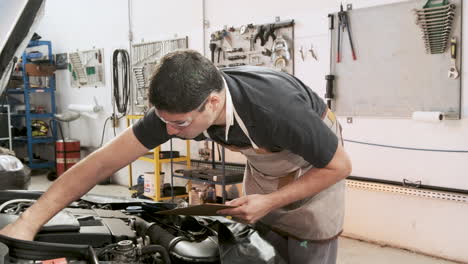 latin adult car mechanic using safety glasses checks the oil level in the car engine, in a repair shop