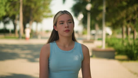 primer plano de una mujer joven con una camiseta de color cian caminando al aire libre, los rayos del sol se reflejan en su mano, con un fondo borroso con árboles, postes de lámparas y vegetación en un brillante día de verano