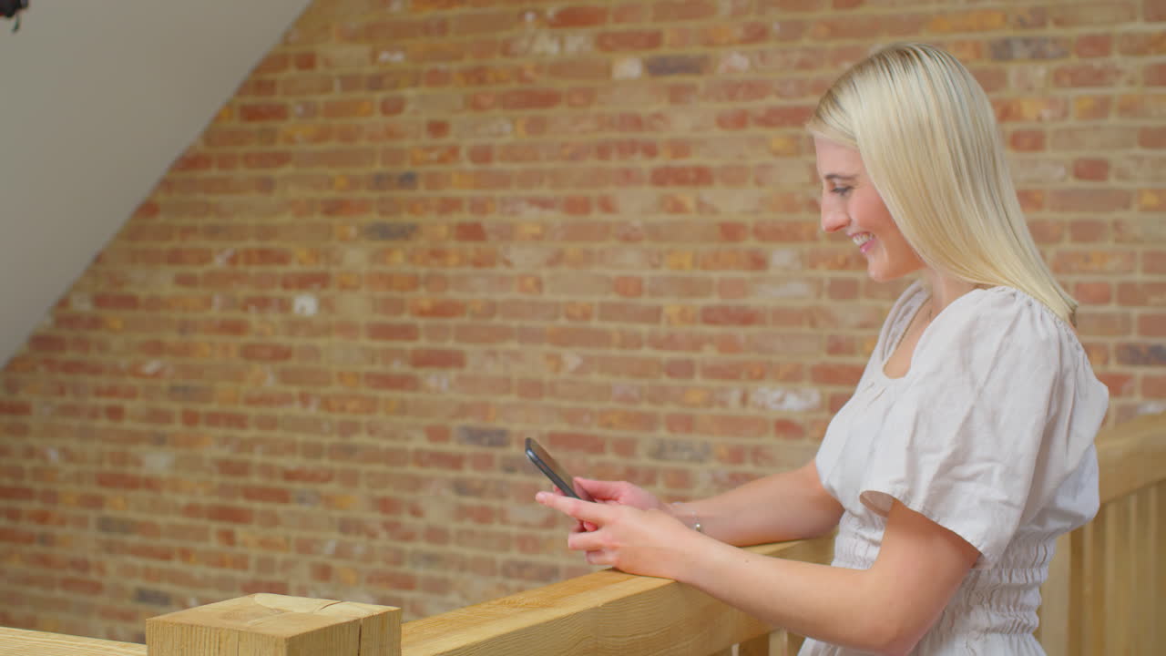 Giovane donna in piedi accanto a una scala di legno a casa che effettua una videochiamata sul cellulare.