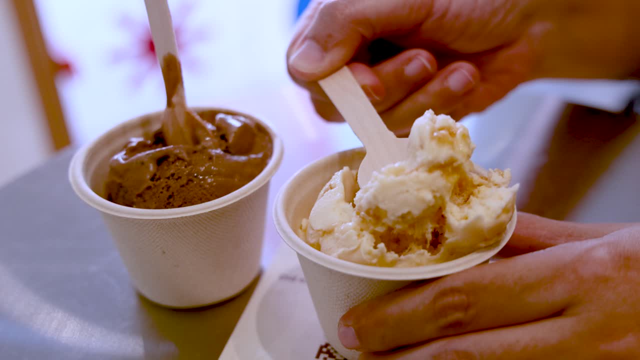 Hand uses wooden spoon to scoop vanilla ice cream from a paper cup beside chocolate ice cream, under soft indoor lighting on a reflective table