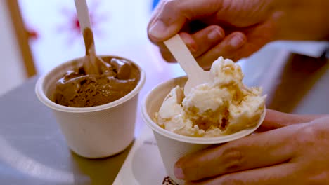 hand uses wooden spoon to scoop vanilla ice cream from a paper cup beside chocolate ice cream, under soft indoor lighting on a reflective table