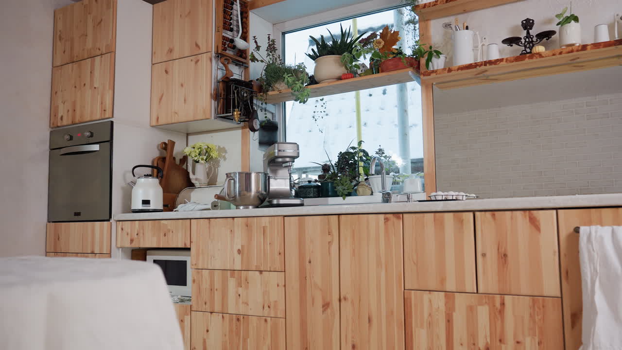 Modern kitchen interior with wooden cabinets, countertop appliances, potted plants, open shelves with kitchenware, and table covered in white cloth near large window