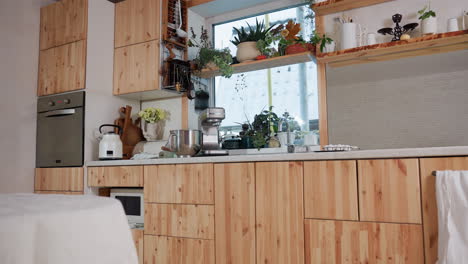 modern kitchen interior with wooden cabinets, countertop appliances, potted plants, open shelves with kitchenware, and table covered in white cloth near large window
