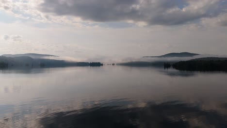 a tranquil lake scene enveloped in morning mist, with soft clouds reflecting on the still waters. the distant islands and surrounding hills create an ethereal and peaceful atmosphere.