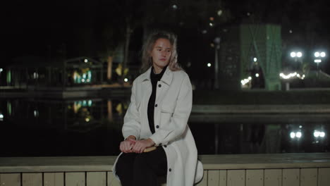 lady in white coat sits on wooden ledge near water at night, ambiance of soft lights reflecting on still surface, capturing relaxed mood in open outdoor environment with mild breeze near city lights