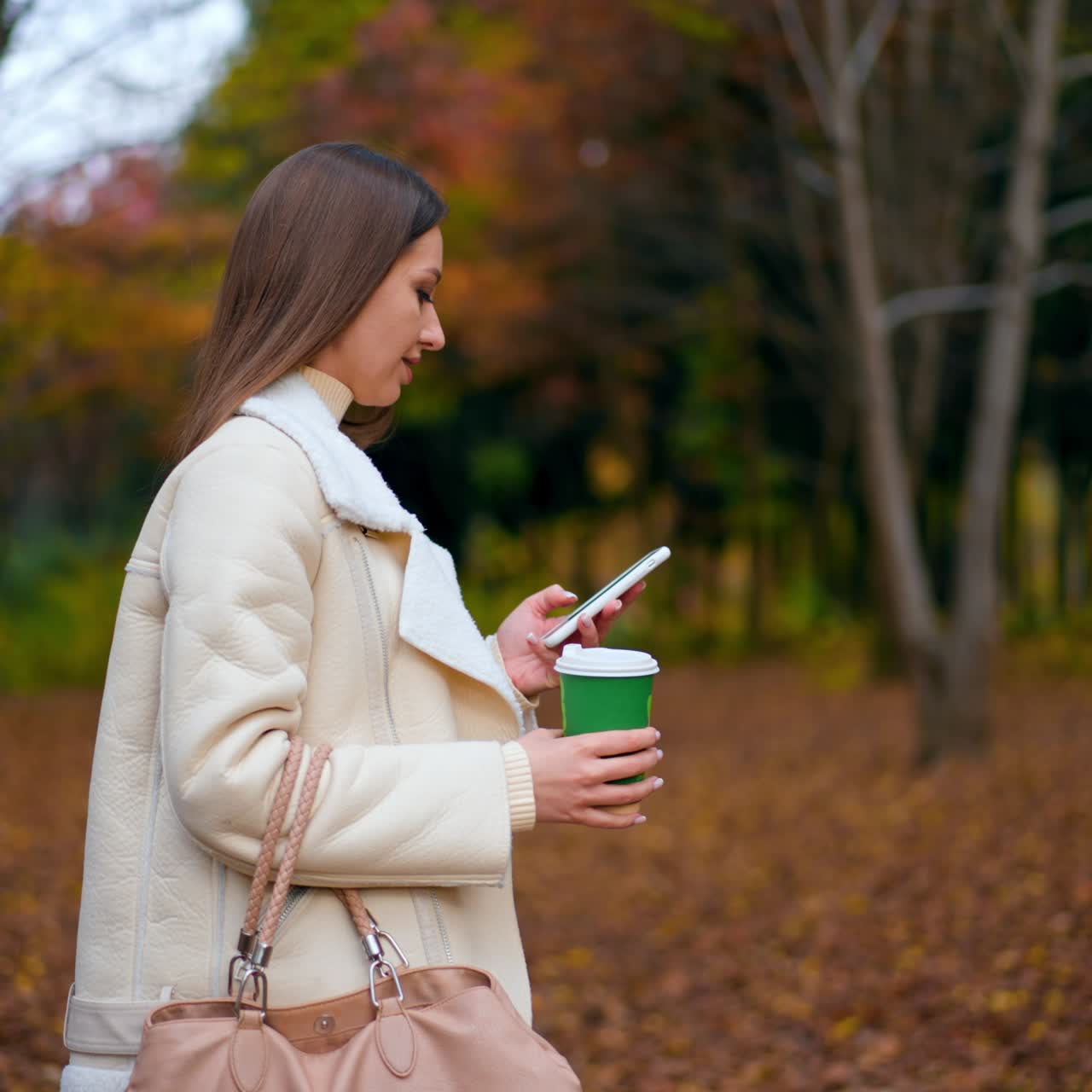 Romantic beautiful lady walking by the autumn park. Woman holding a paper cup in her hand and looking at smartphone. Blurred backdrop