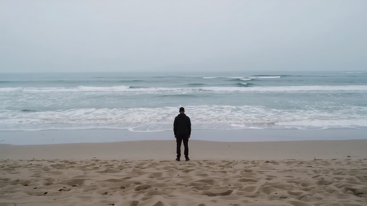Uomo in piedi con giacca e berretto, di fronte all'oceano in spiaggia, che osserva le onde che si avvicinano e si ritirano.