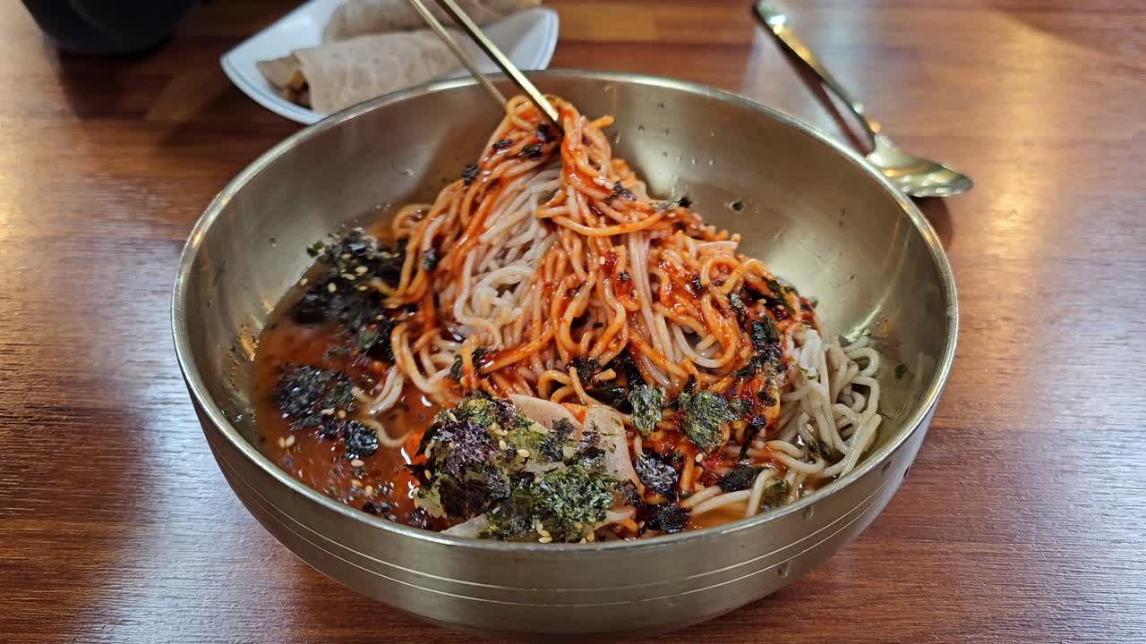 Le bacchette mescolano spaghetti di grano saraceno piccanti rossi al gochujang, guarniti con alghe essiccate e semi di sesamo, in una tradizionale ciotola d'acciaio coreana, serviti con involtini bossam.